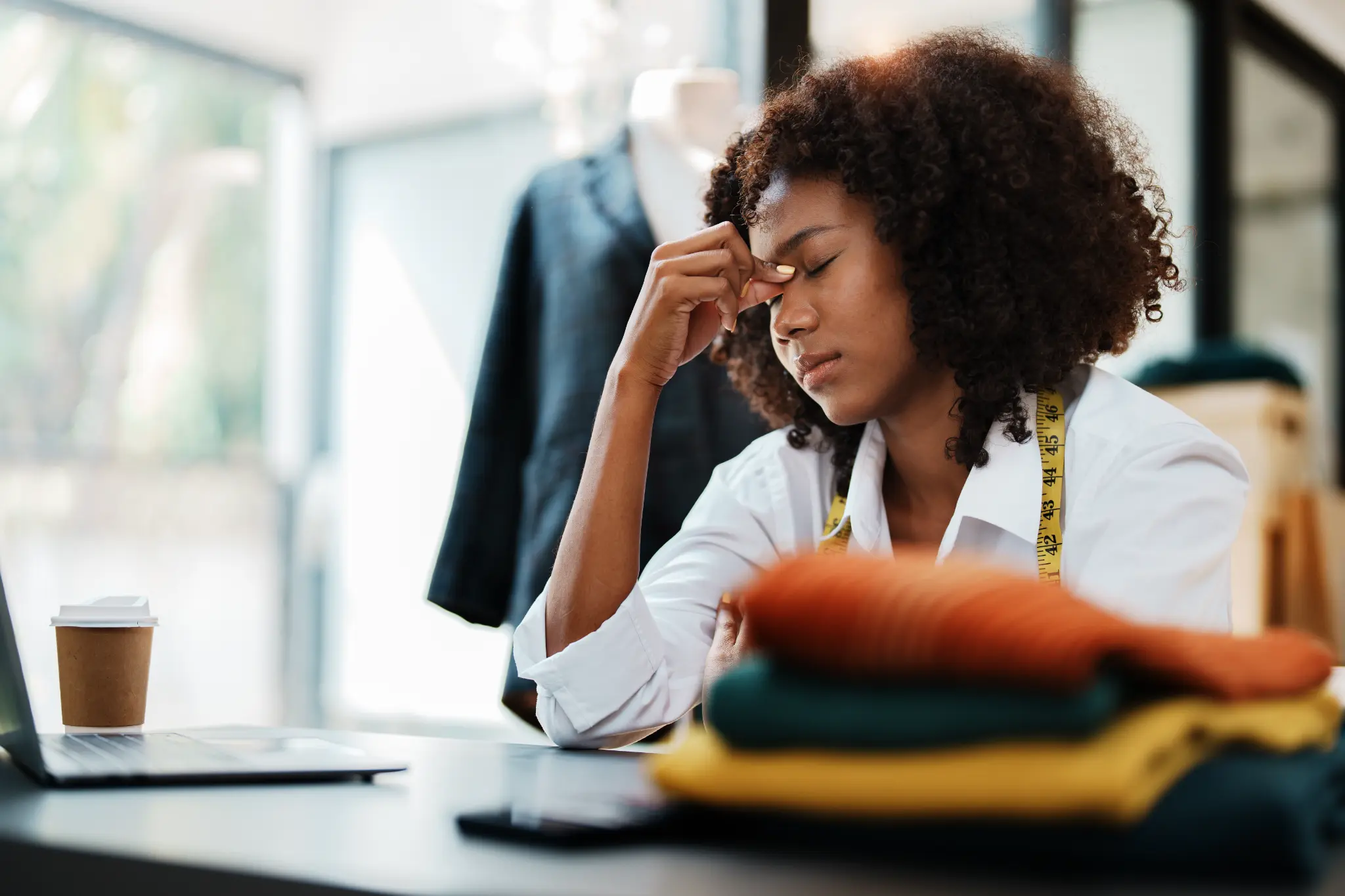 Fashion designer pausing at her worktable beside folded fabrics and a laptop